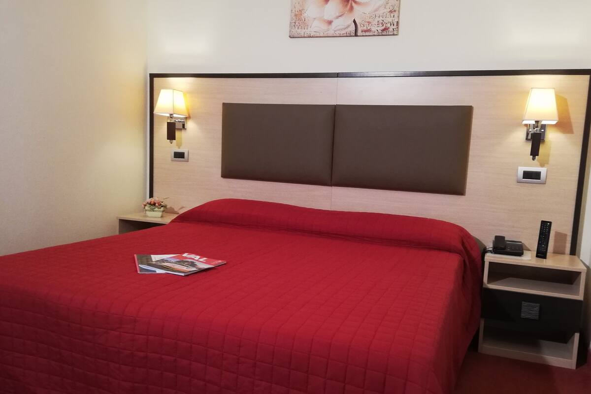 A hotel room with a large bed, two bedside tables, a nightstand, a magazine on the bed, and two wall-mounted lamps.