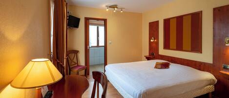 Habitación de hotel con cama, escritorio con lámpara, silla, televisor y puerta que da al baño.