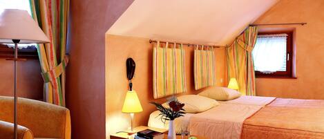 Quarto de hotel com uma cama, duas cadeiras, uma mesa de cabeceira com abajur, um vaso com flores e uma janela com cortinas.