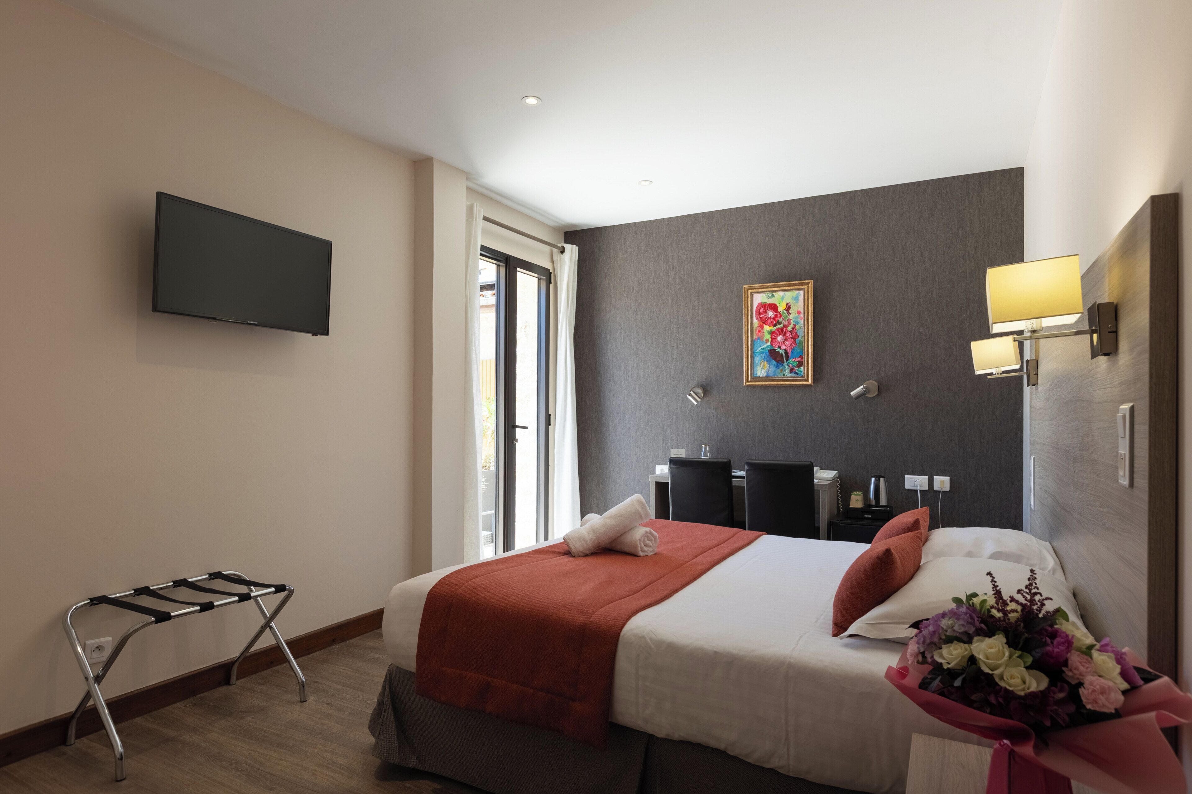 A hotel room with a bed, a flat-screen TV, a painting, a desk with a lamp, and a bouquet of flowers.