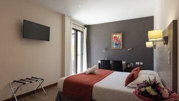 A hotel room with a bed, a flat-screen TV, a painting, a desk with a lamp, and a bouquet of flowers.