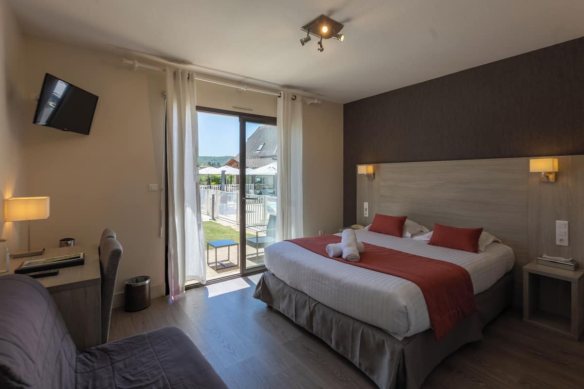 Ein Hotelzimmer mit Bett, Fernseher, Schreibtisch, Stuhl, Lampe und Blick ins Freie.
