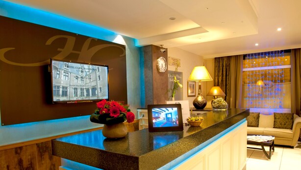 Zona de recepción de un hotel con un mostrador, un reloj, un televisor mostrando información del hotel, un jarrón con flores y una ventana con luces azules.