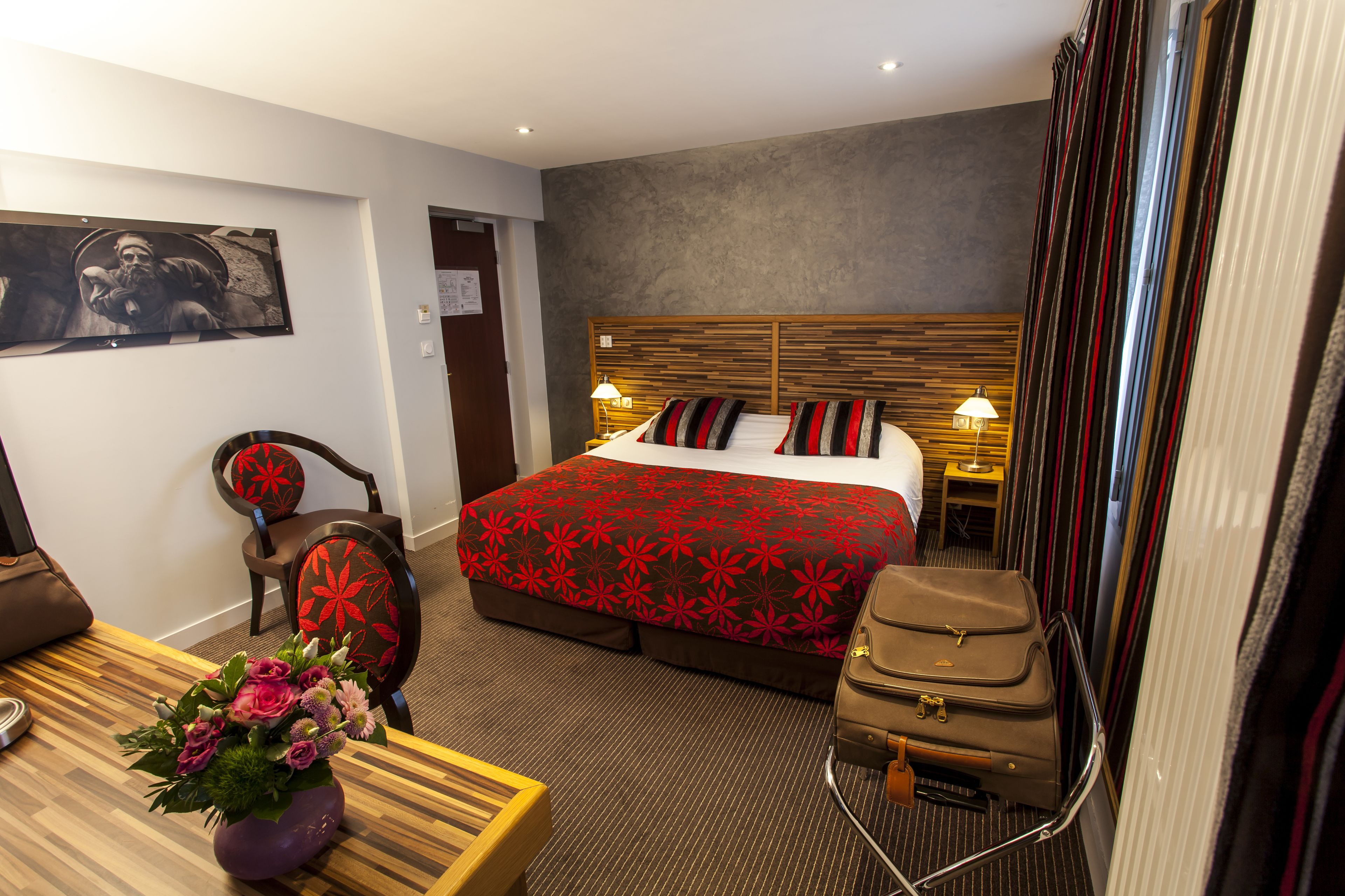 Una habitación de hotel con cama, escritorio, silla, mesita de noche, lámpara, maleta y un jarrón con flores.