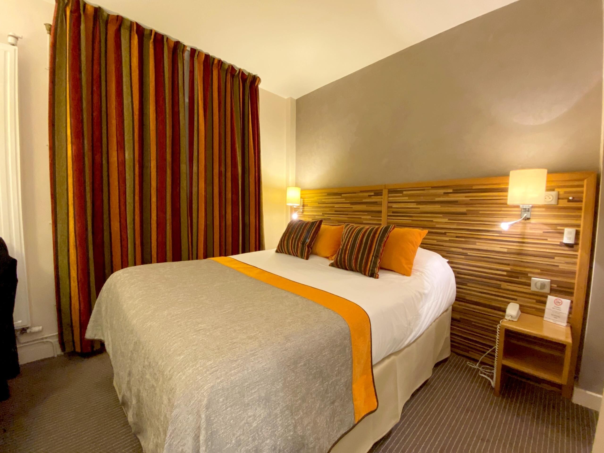 A hotel room with a large bed, striped curtains, a wooden headboard, bedside lamps, and a small bedside table.