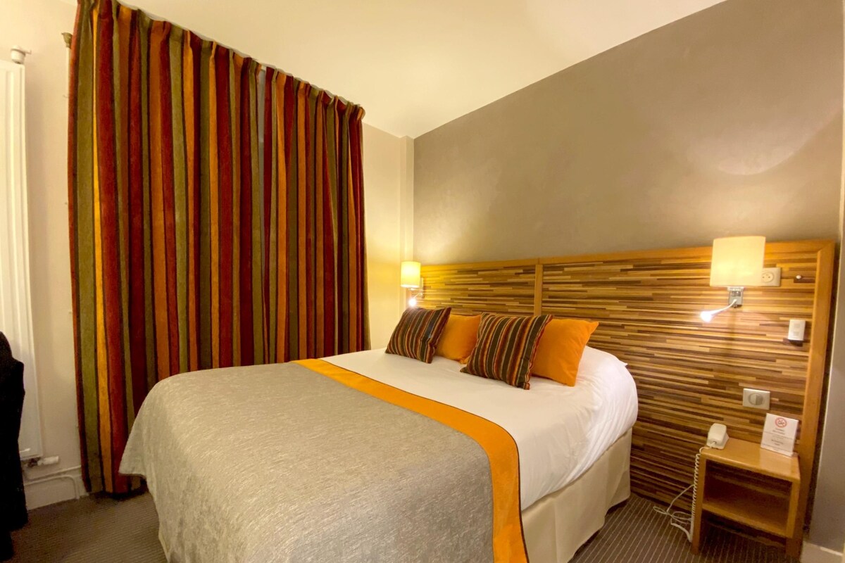 A hotel room with a large bed, striped curtains, a wooden headboard, bedside lamps, and a small bedside table.