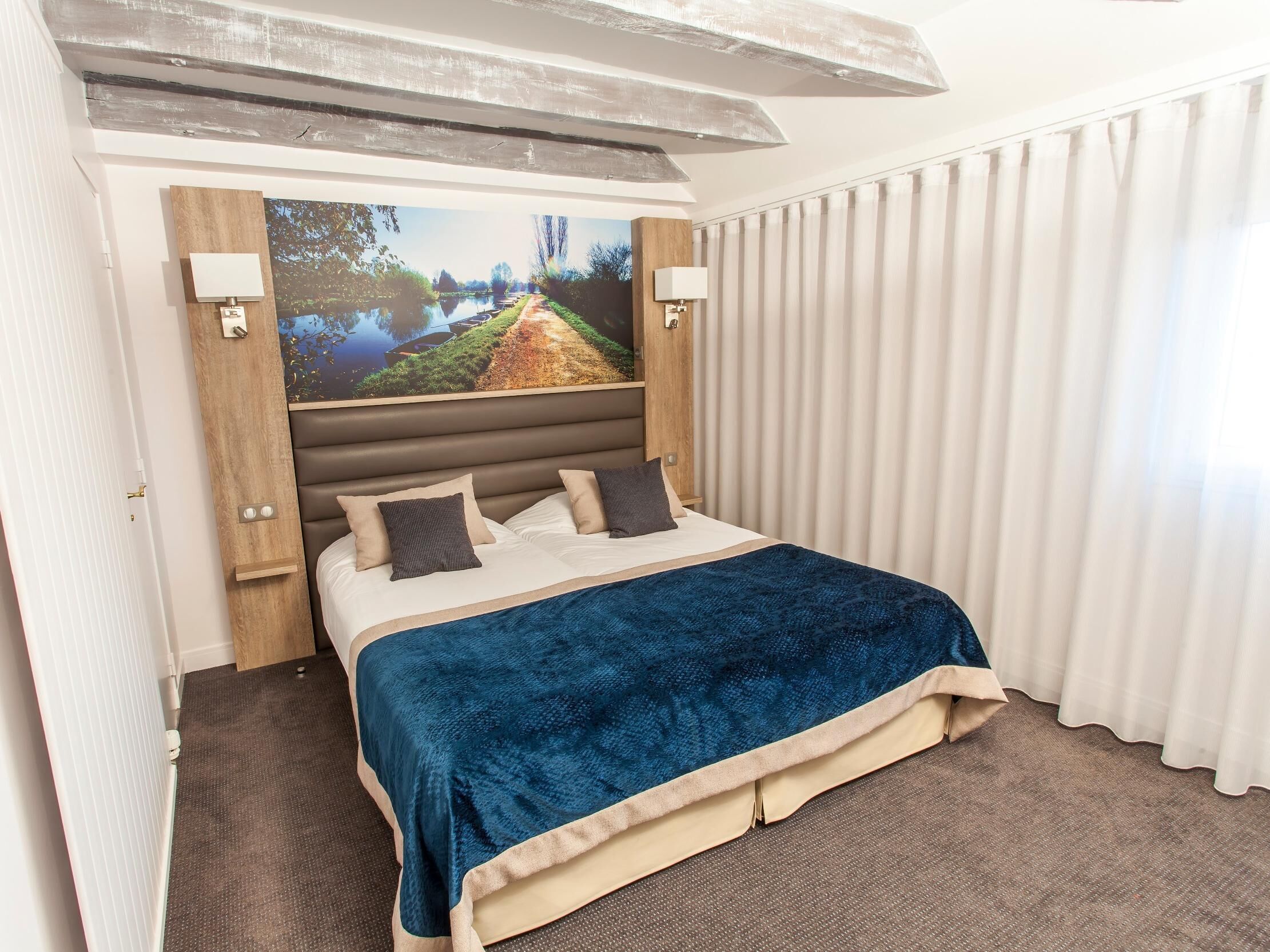 A hotel room with a large bed, a headboard with a painting, two bedside lamps, and a view of a river through the window.