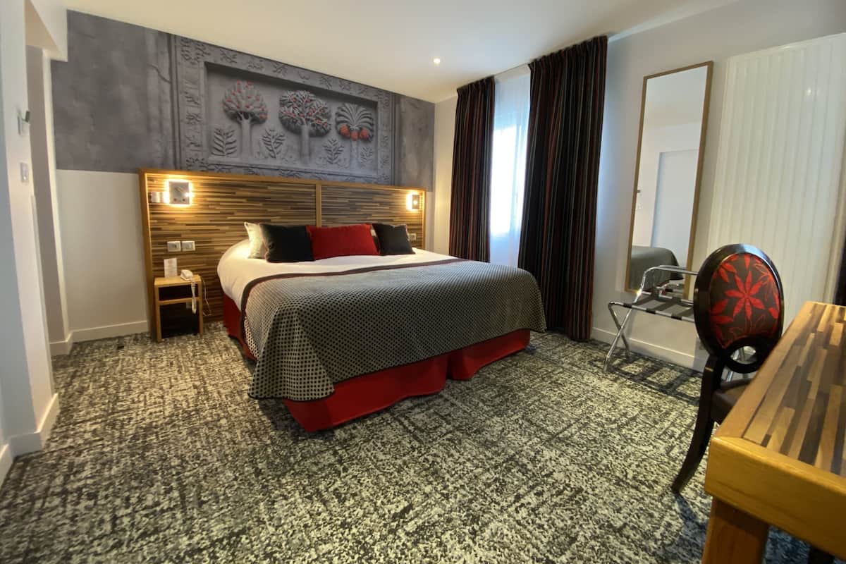 A hotel room with a large bed, a wooden headboard, a small bedside table with a phone, a desk with a chair, and a window with curtains.