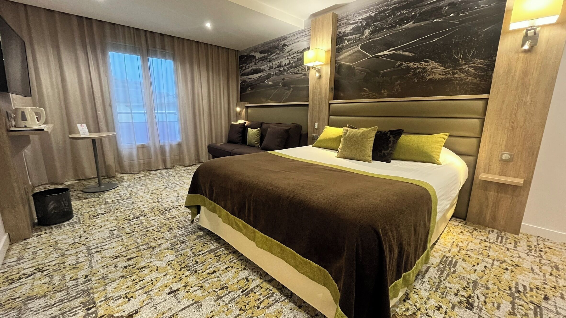 A hotel room with a large bed, a headboard with a landscape mural, a small table with a kettle, and a view of the outdoors through the window.