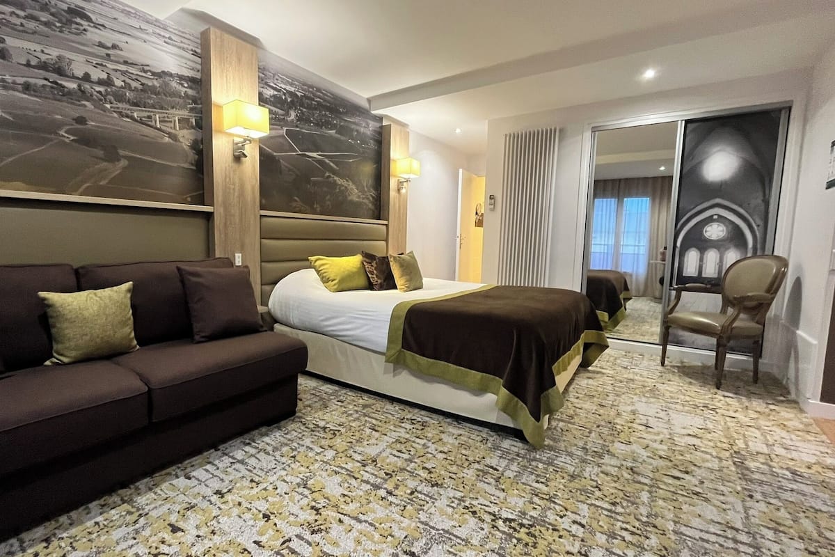 A hotel room with a bed, a dark-colored sofa, a patterned carpet, a large wall mural, and a chair.