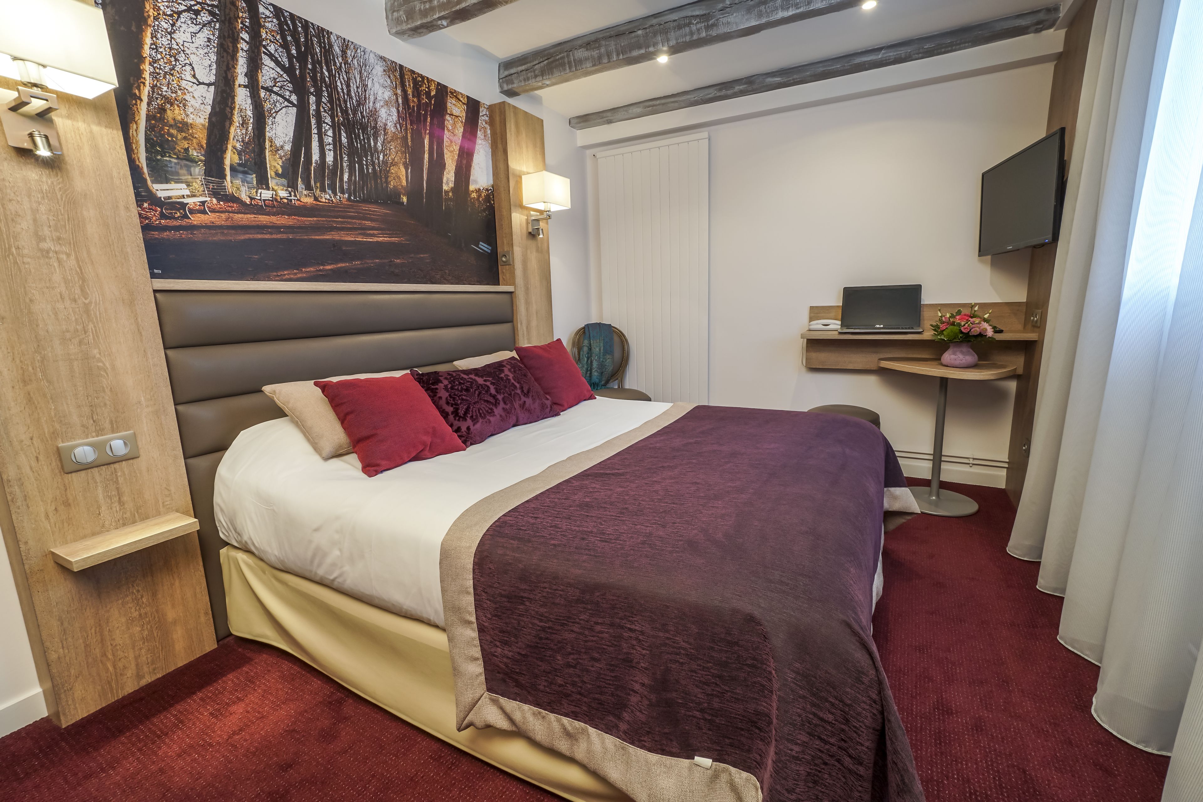 A bedroom with a large bed, a desk with a laptop, a TV, and a painting of a forest.