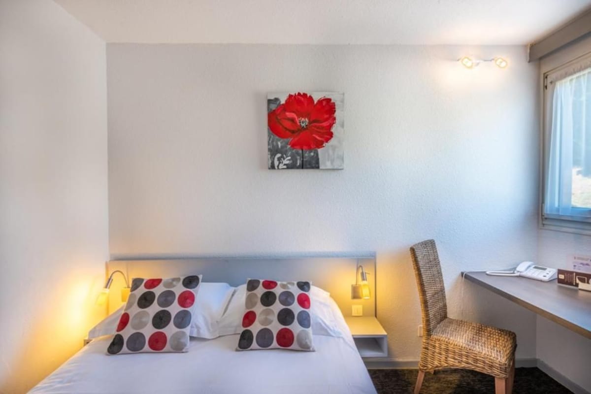 A hotel room with a bed, a chair, a desk, a phone, and a painting on the wall.