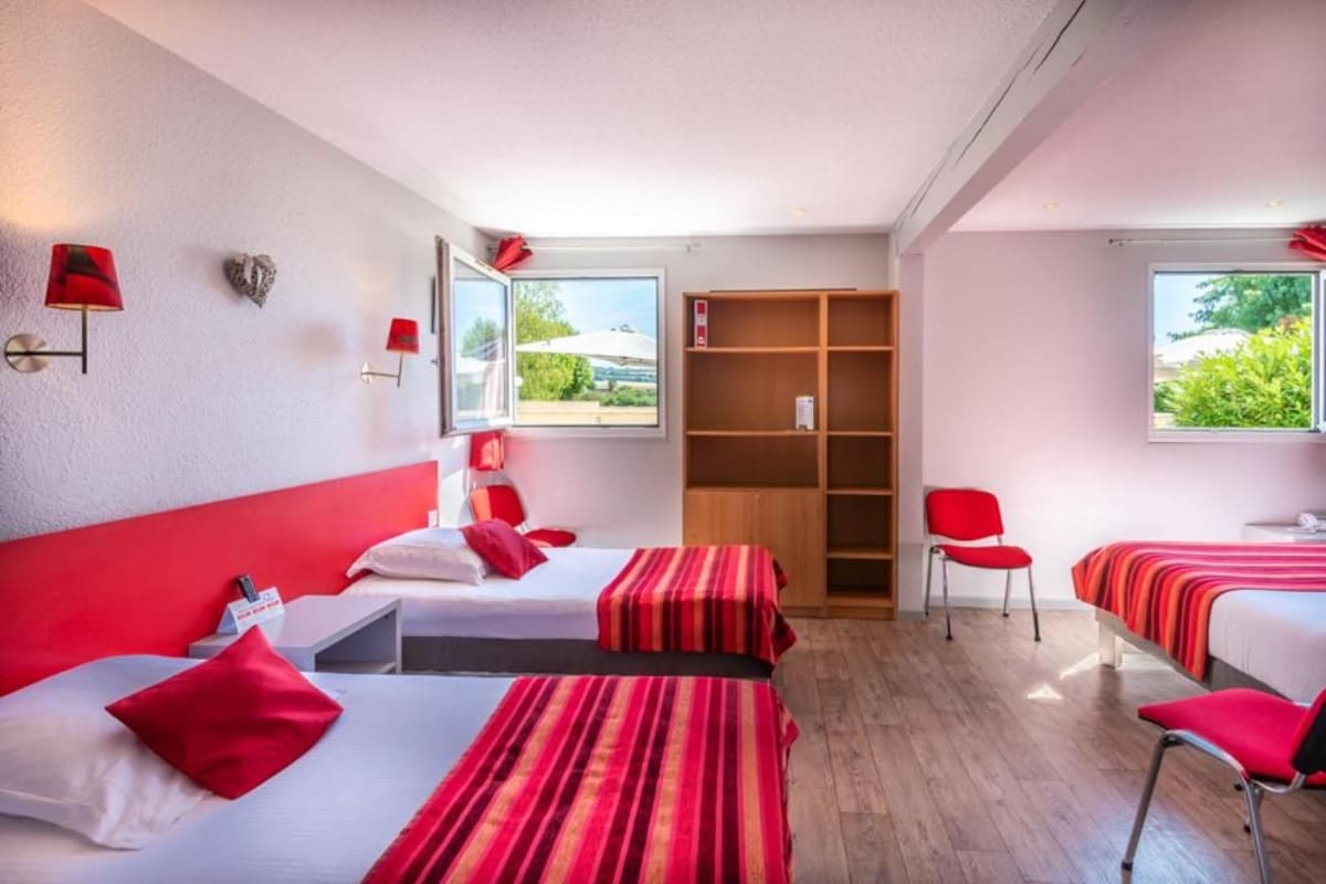A hotel room with two beds, a wooden bookshelf, a red chair, and a window with a view of greenery.