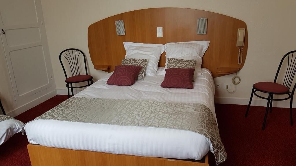 A hotel room with a wooden headboard, a bed with patterned bedding, two chairs, and a telephone.