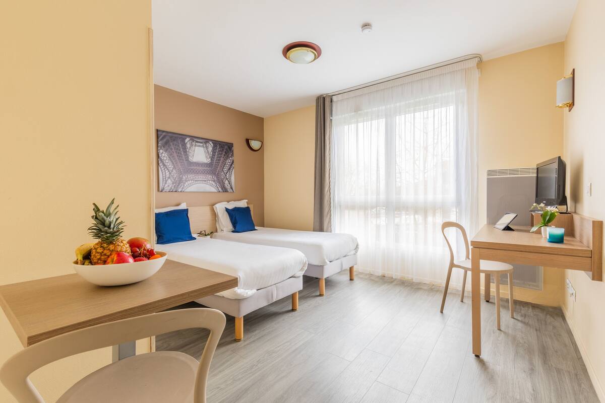 A hotel room with two beds, a desk, a chair, a TV, and a fruit bowl.