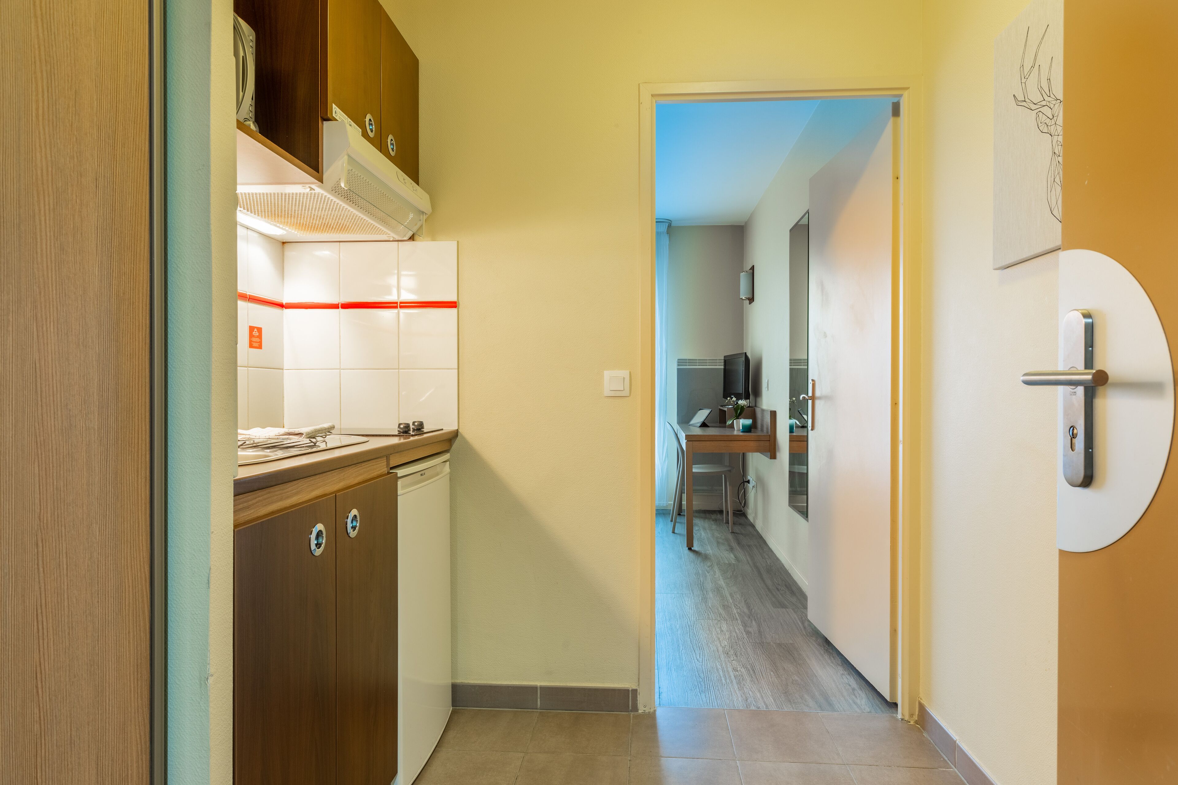 Un couloir étroit, avec un coin cuisine sur la gauche, comprenant une cuisinière, des placards et un pare-feu en carrelage. On aperçoit une table à manger et des chaises au fond.