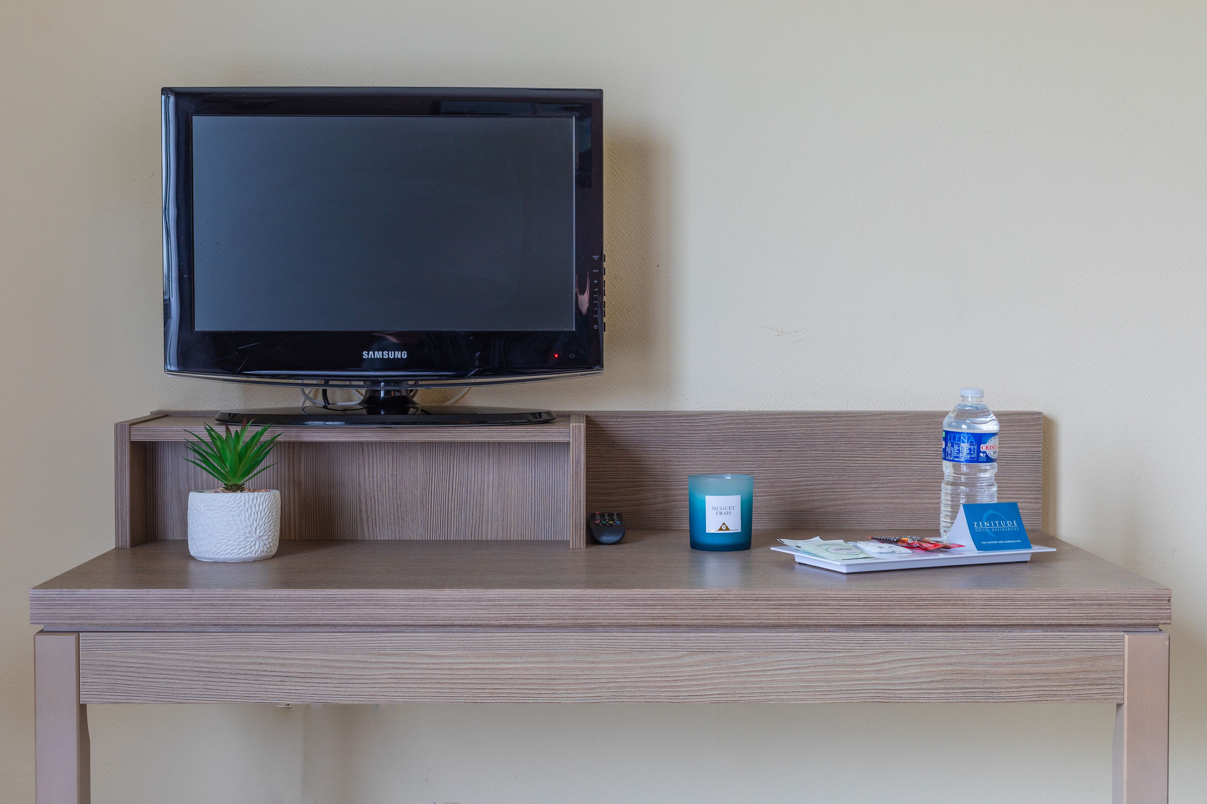 Quarto de hotel com televisão de tela plana, uma pequena planta em vaso, uma garrafa de água, uma vela e um tabuleiro com cartão e caneta.