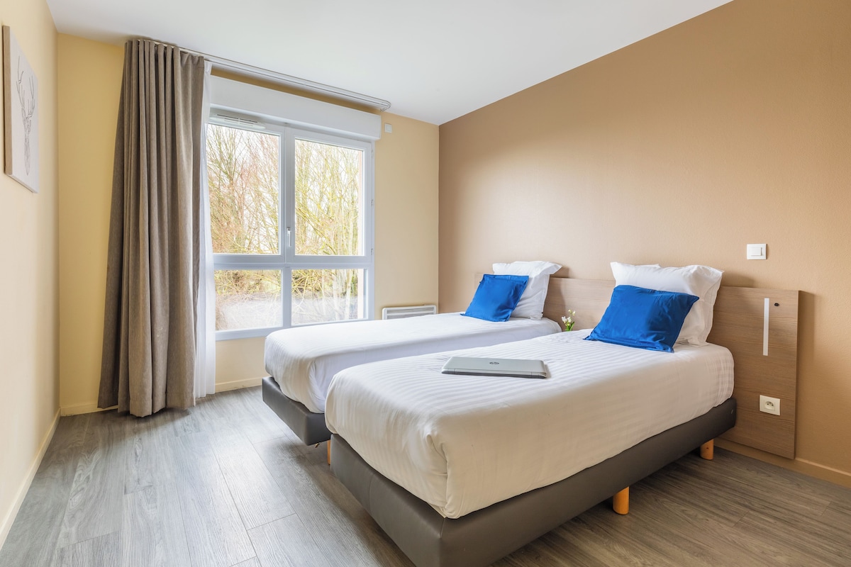 Quarto de hotel moderno com duas camas, uma janela grande e vista para árvores.