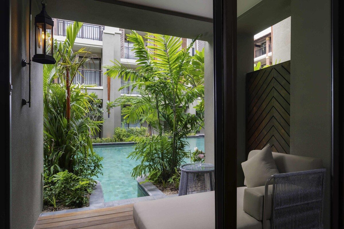 A balcony with a pool, surrounded by lush greenery and a view of a building with balconies.