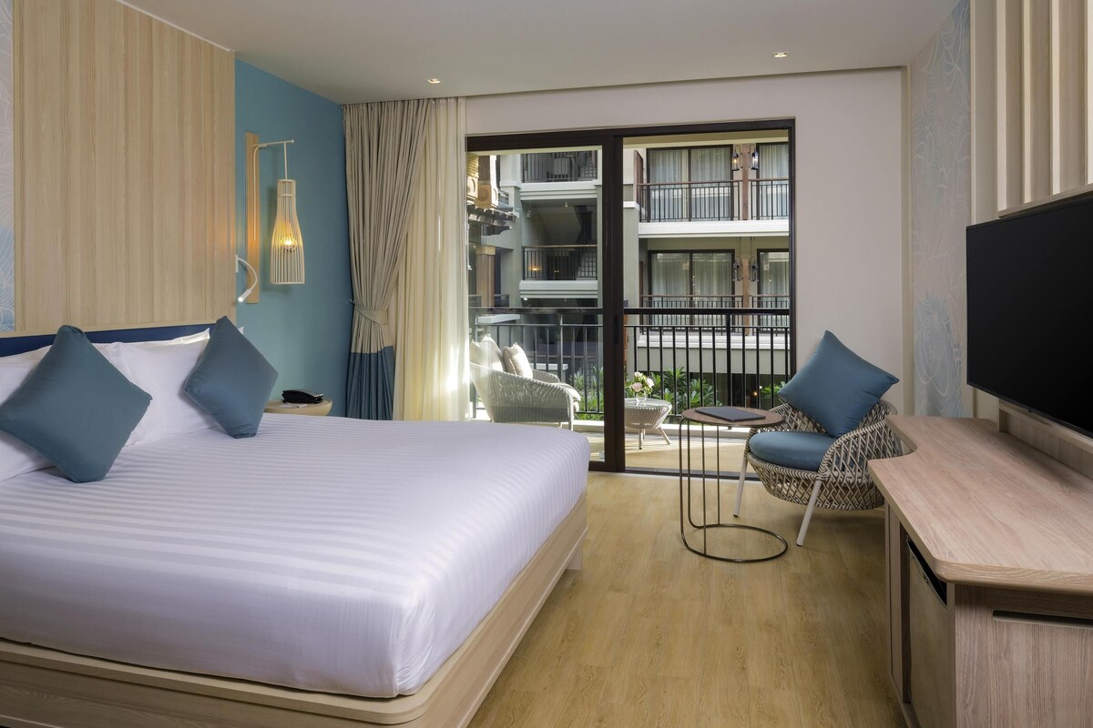 A hotel room with a bed, a TV, a desk, a chair, a balcony with a view of the building, and a window with curtains.