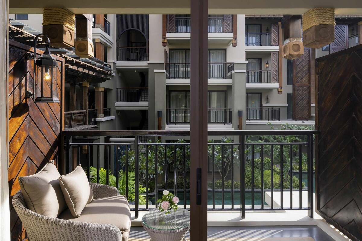 A balcony with a wicker sofa, a glass table with flowers, and a view of a pool and greenery.
