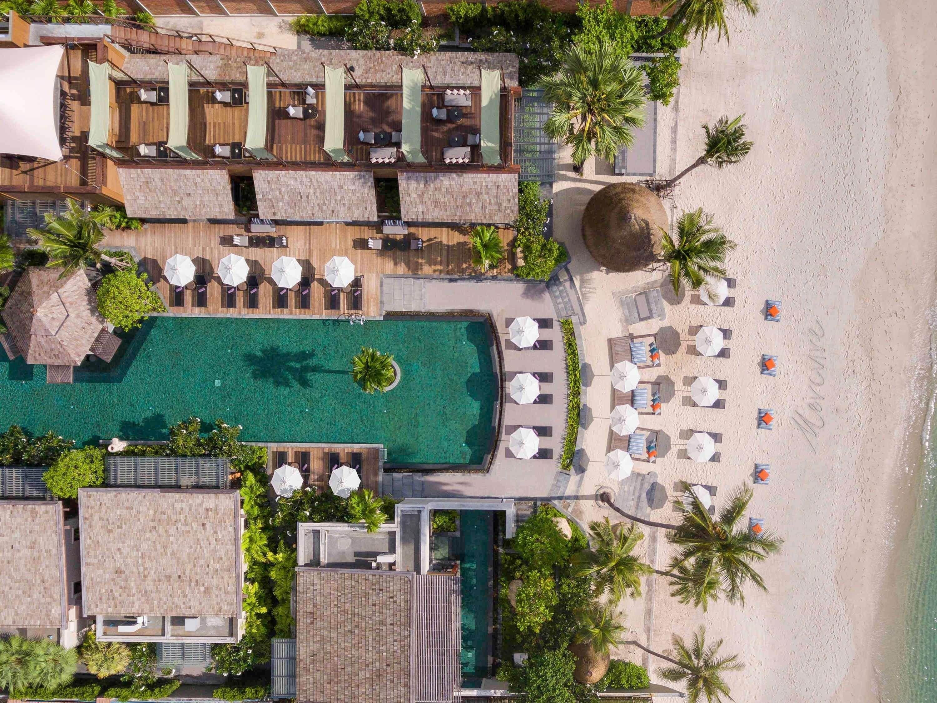 Vista aérea de un resort con una piscina central, varias zonas de descanso con sombrillas y un área de playa con tumbonas.
