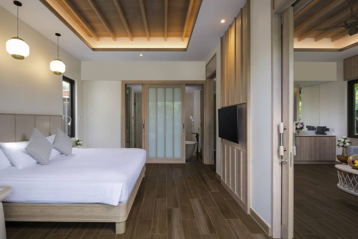 A modern hotel room with a large bed, wooden flooring, a TV, and a bathroom visible through a sliding door.