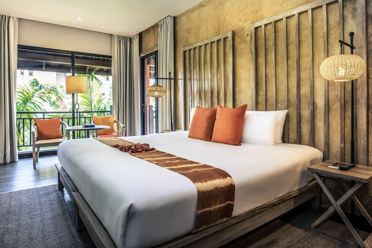 A hotel room with a large bed, a wooden headboard, a small table with a lamp, and a view of greenery outside.