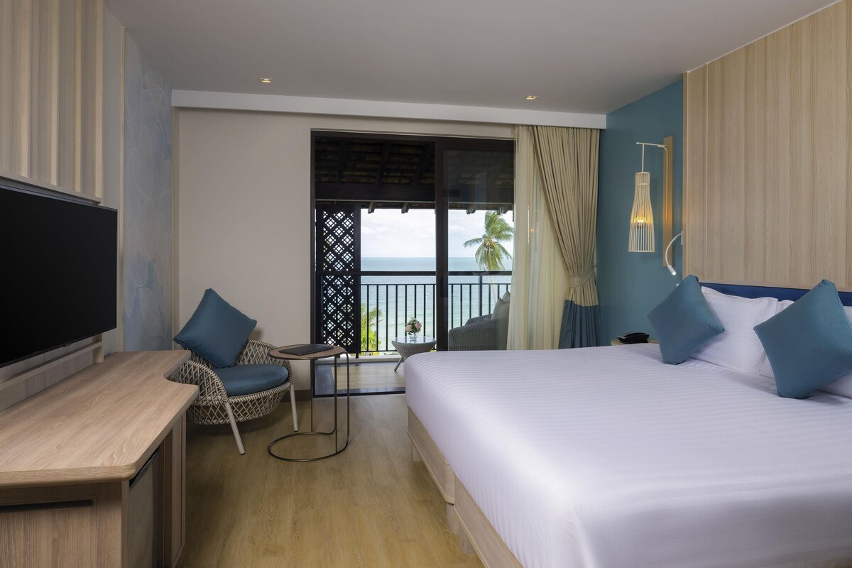 A hotel room with a bed, a desk, a chair, a TV, and a balcony with a view of the ocean.