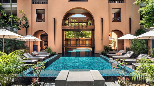 Una piscina con sillas de descanso y sombrillas, rodeada de plantas y un edificio con balcones.