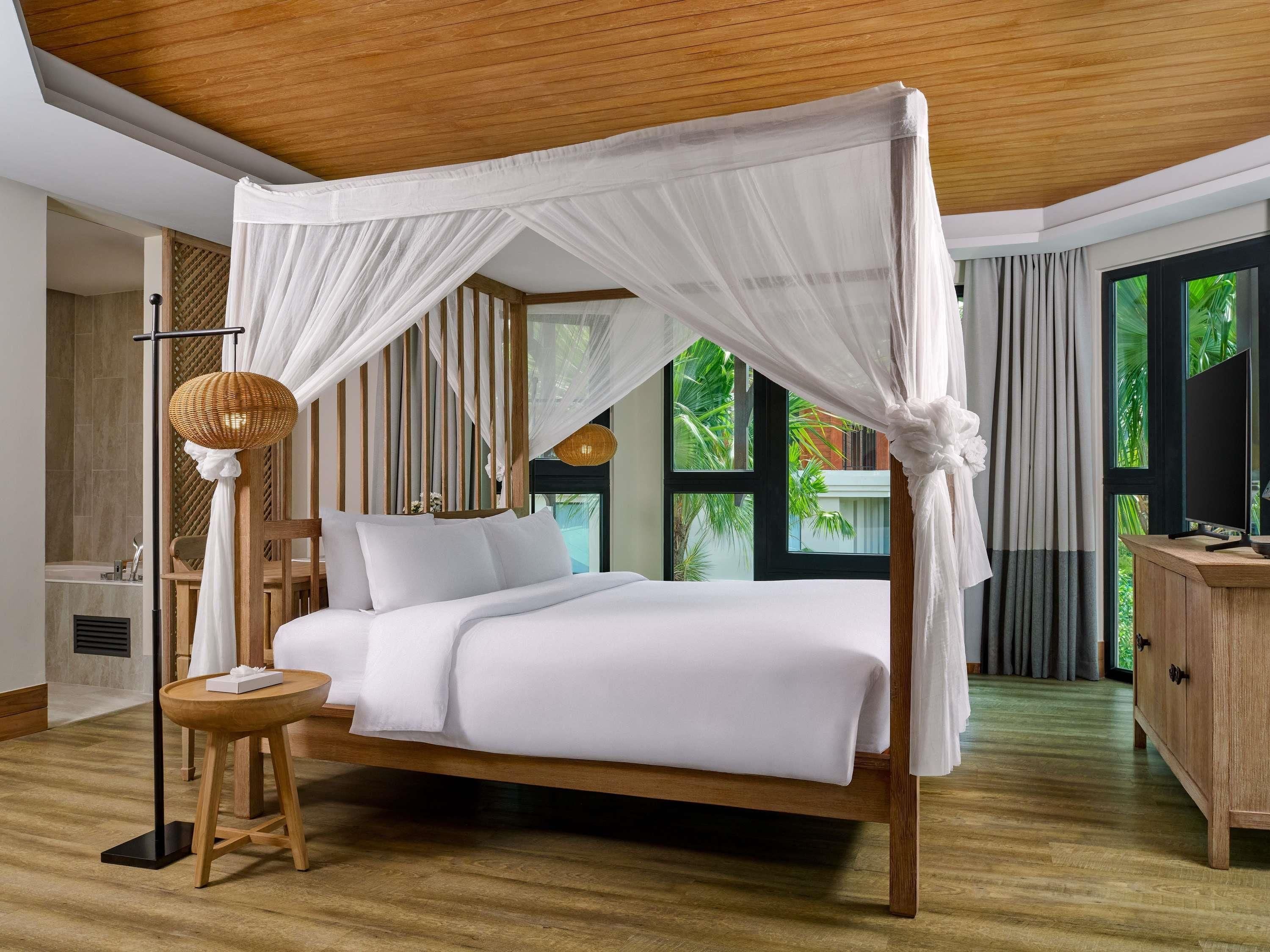 A bedroom with a canopy bed, a bedside table, a lamp, and a view of greenery outside.