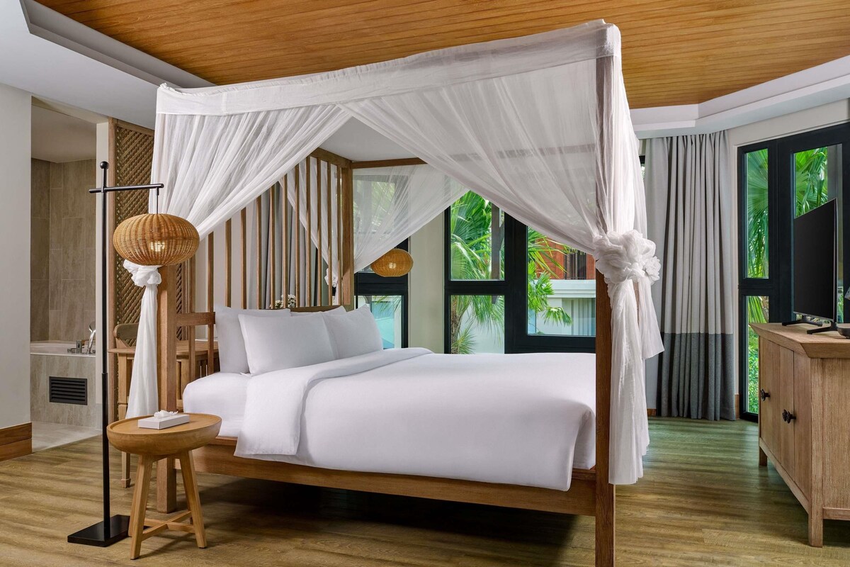 Un dormitorio con cama de dosel, una mesita de noche, una lámpara y vistas al verdor exterior.