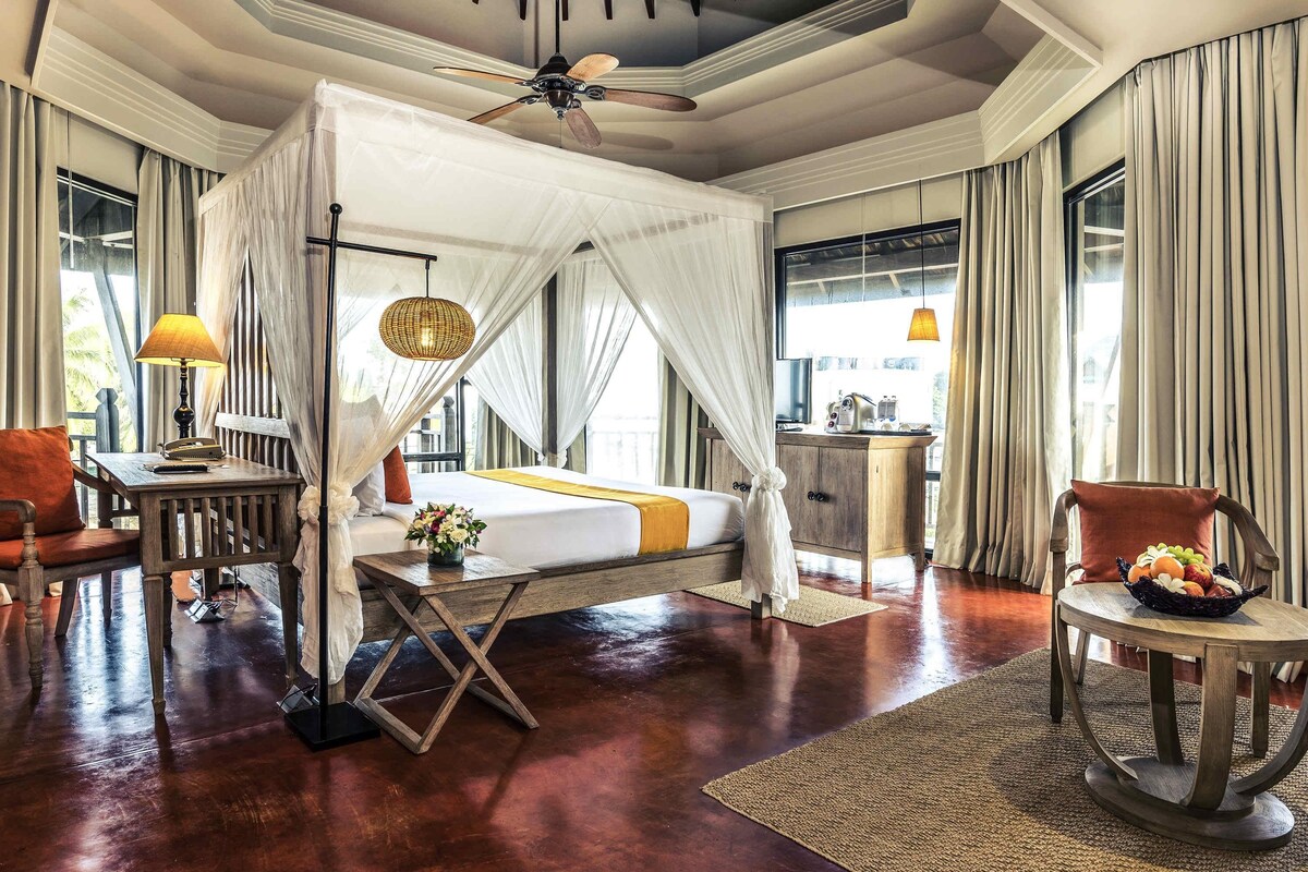 A spacious room with a large bed, a ceiling fan, a desk with a lamp, and a small table with fruit.