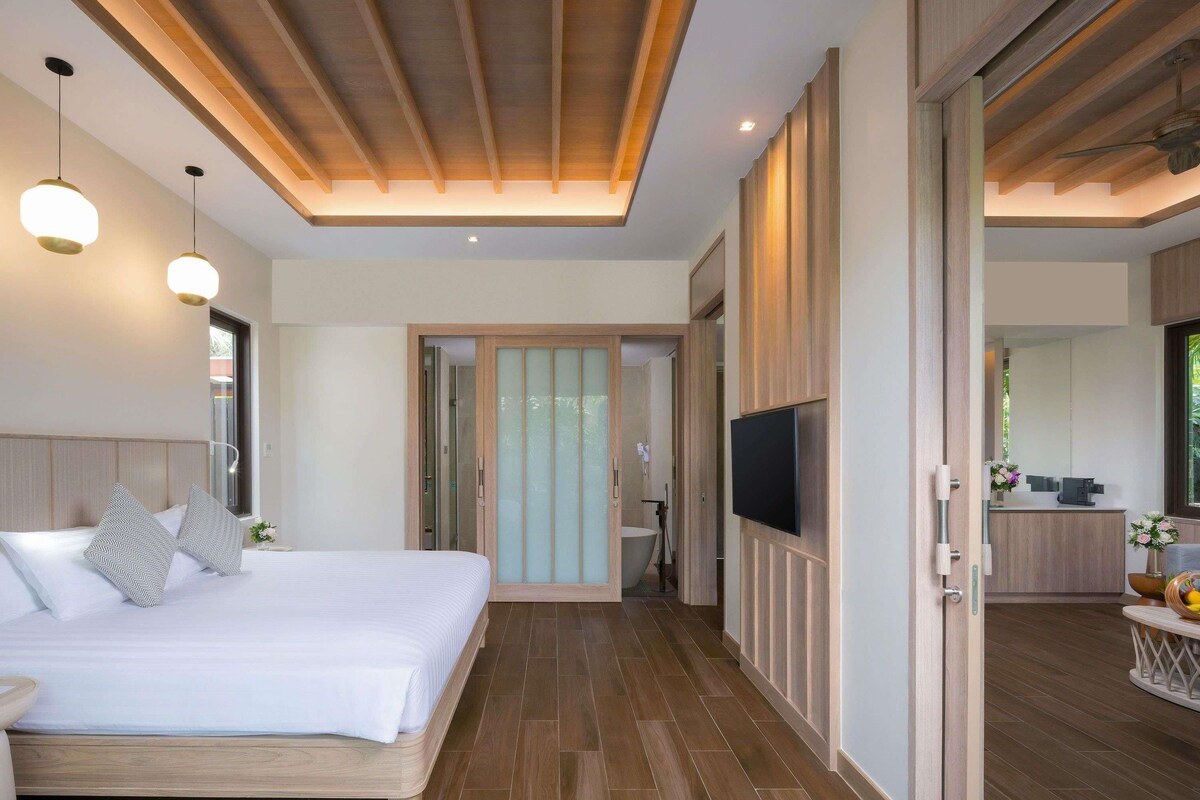 A modern hotel room with a wooden bed, a TV mounted on the wall, and a bathroom visible through an open door.