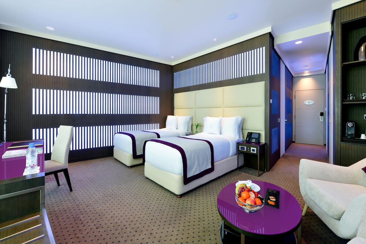 A hotel room with two beds, a desk, a chair, a small table with fruit, and a minibar.