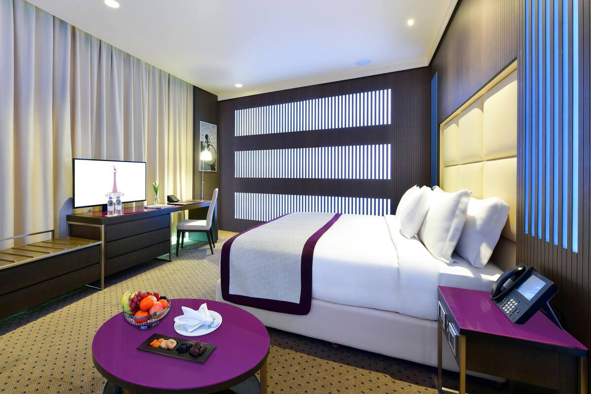 A modern hotel room with a large bed, a desk, a chair, a small table with fruit, and a TV.