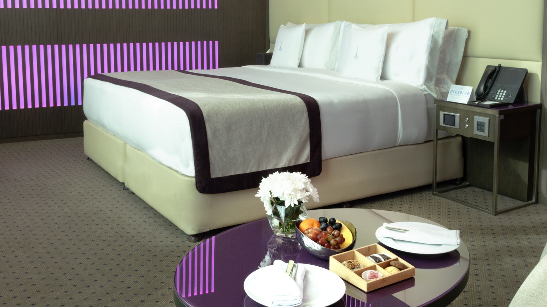 Hotelkamer met een bed, nachtkastje en een tafeltje met een bloevas, schaal fruit en een bord.