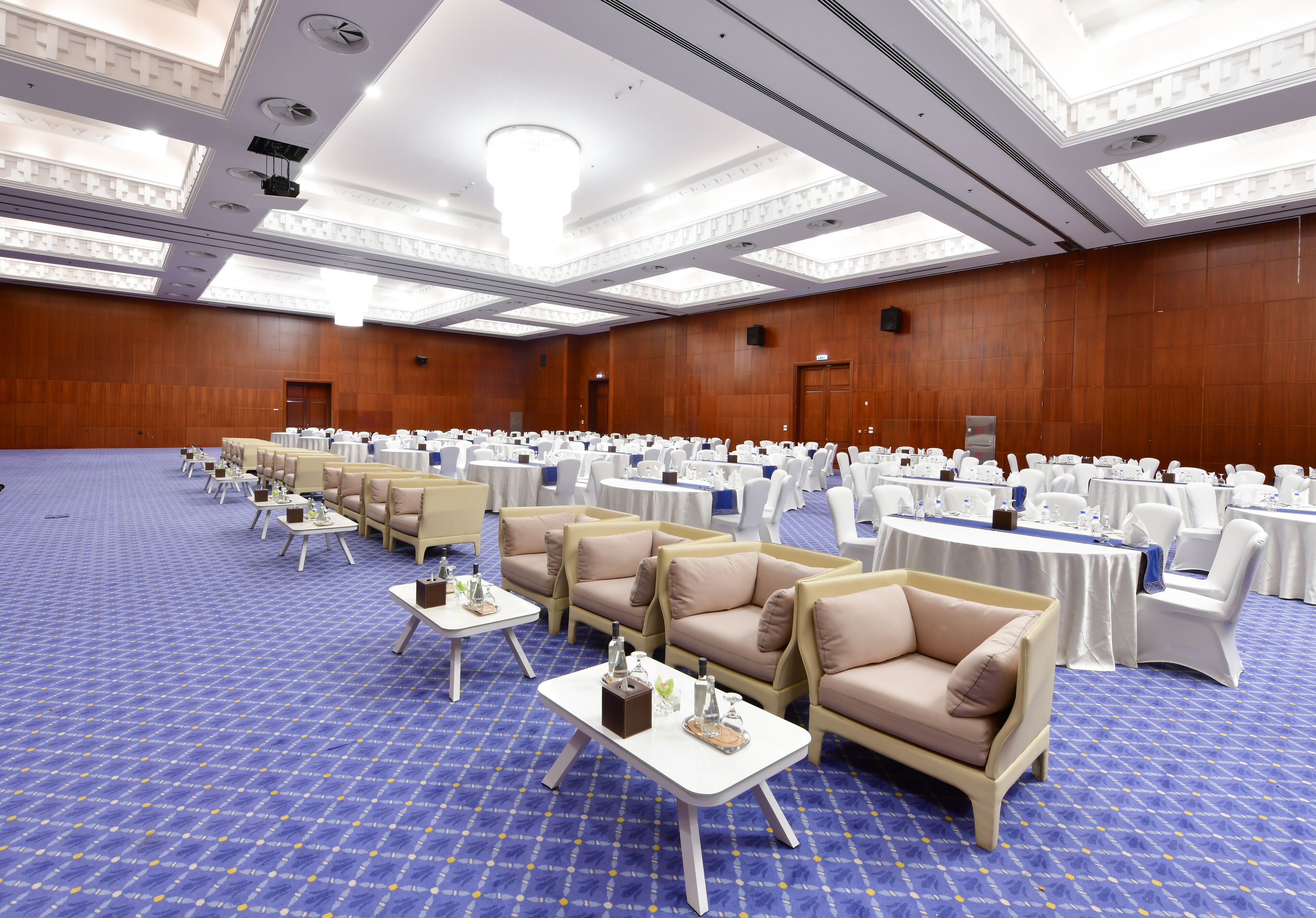 A large conference room with rows of white tables and chairs, beige sofas, and a blue carpet.