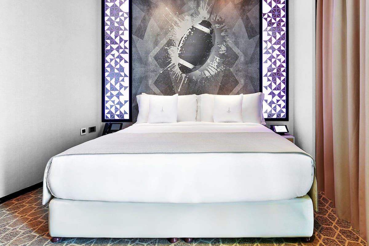 A double bed with white bedding, a large abstract artwork above it, and patterned curtains.
