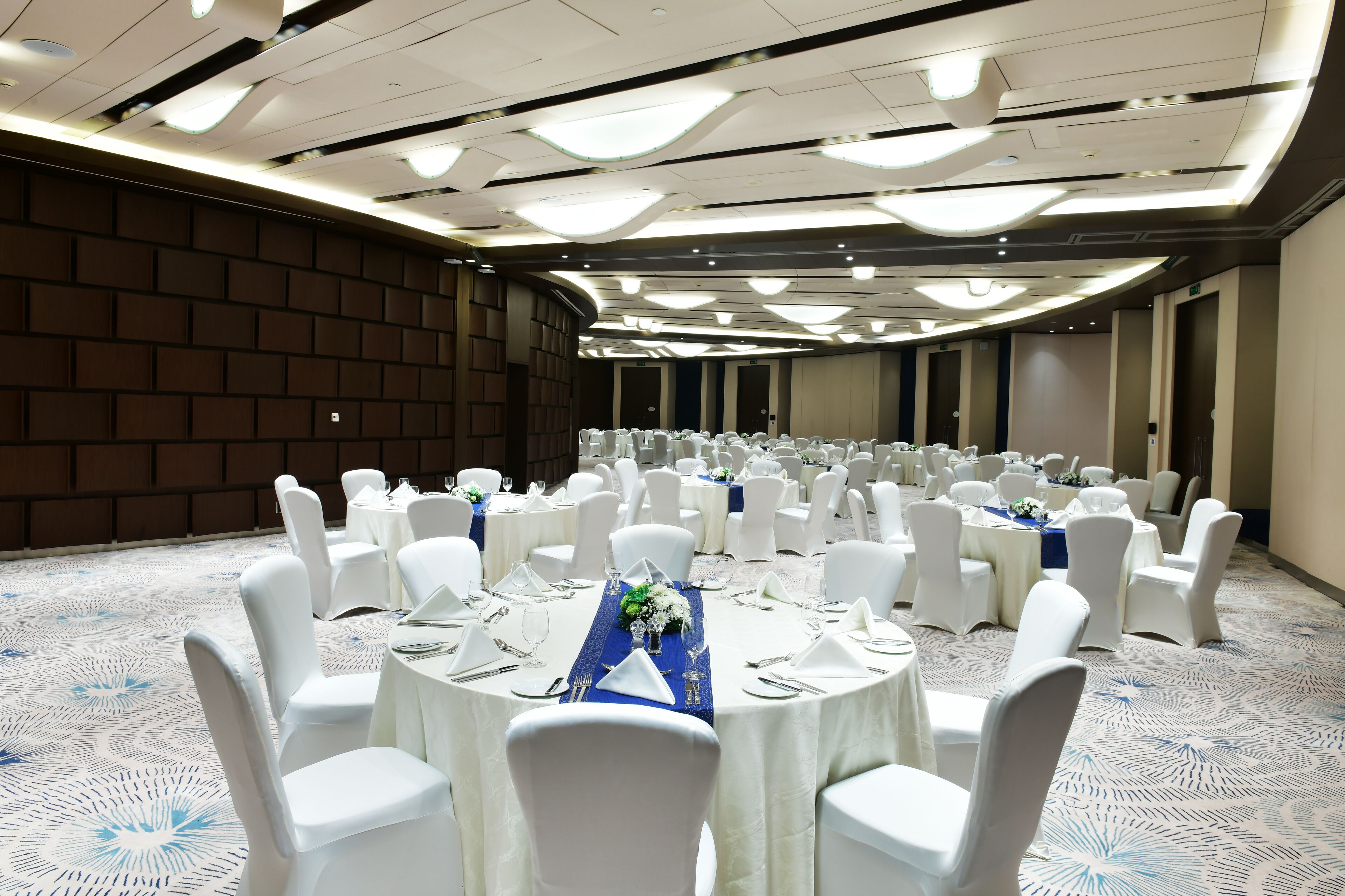 Une grande salle de banquet avec des tables rondes, des chaises blanches et un tapis à motifs.