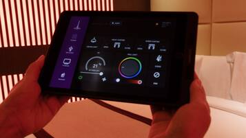 Una tableta muestra la interfaz de control de iluminación de la habitación, con opciones para la luz del techo, la luz de pared y la configuración de las cortinas.
