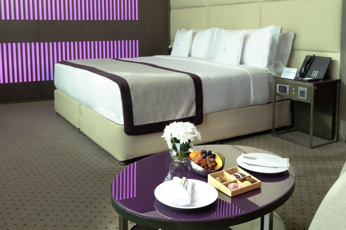 A hotel room with a large bed, a bedside table with a phone, a small table with a vase of flowers, and a fruit bowl.