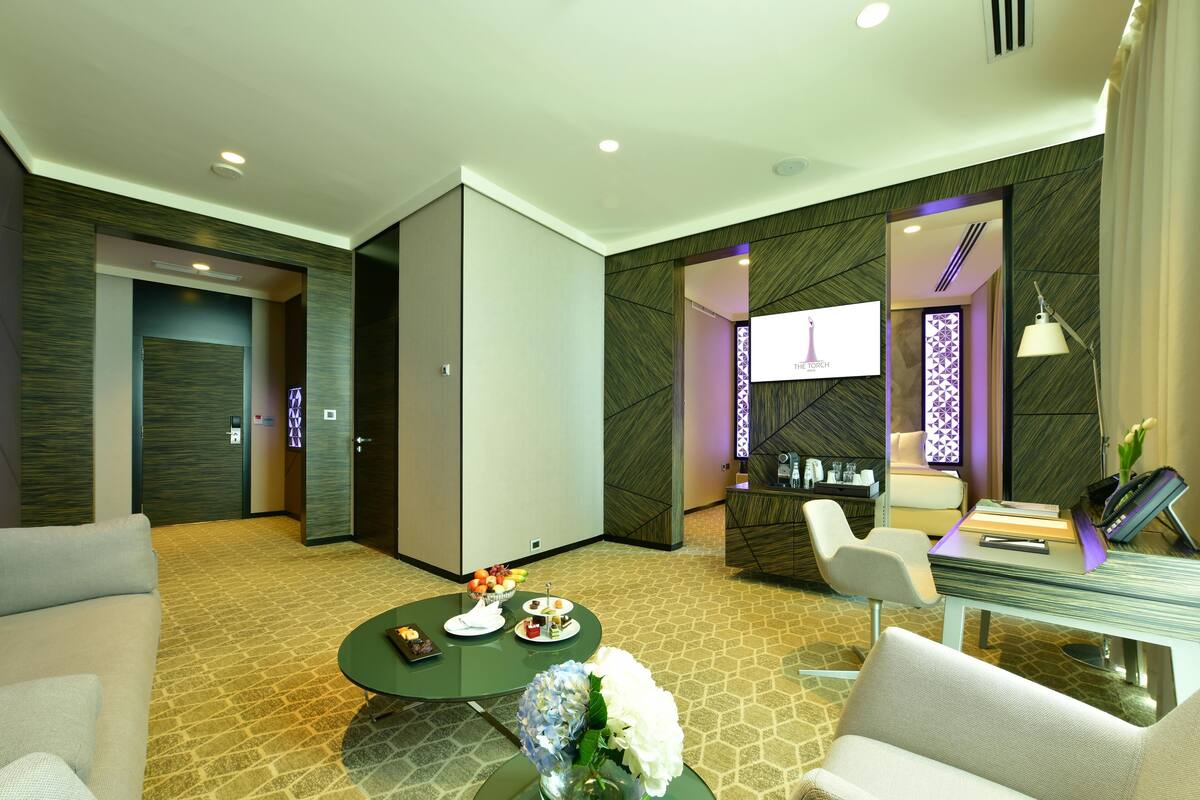 A modern hotel room with a sofa, a round table with a vase of flowers, a desk with a lamp, and a TV on the wall.