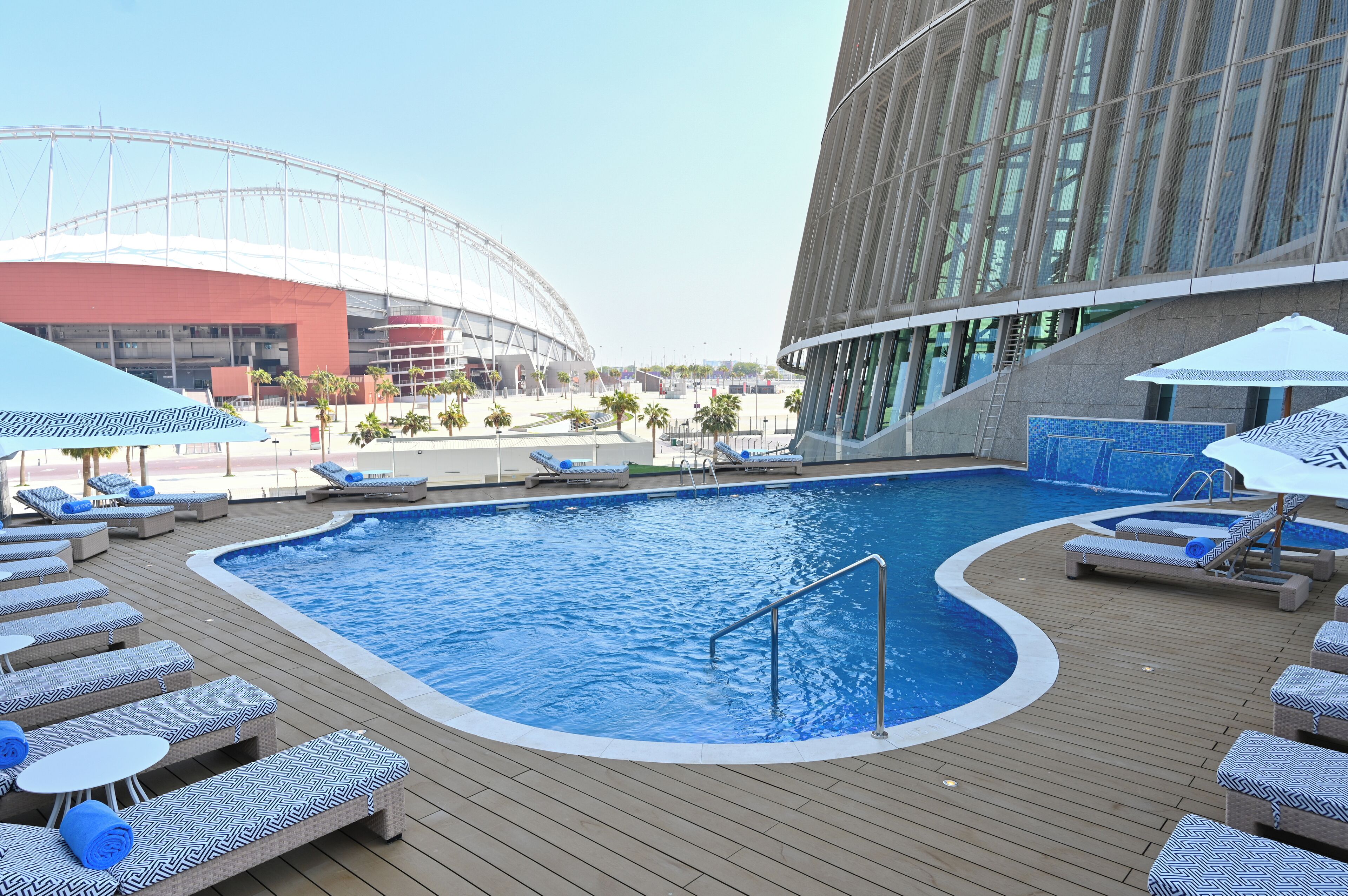 Una piscina con sillas de descanso y sombrillas, un edificio moderno con grandes ventanales y un estadio al fondo.