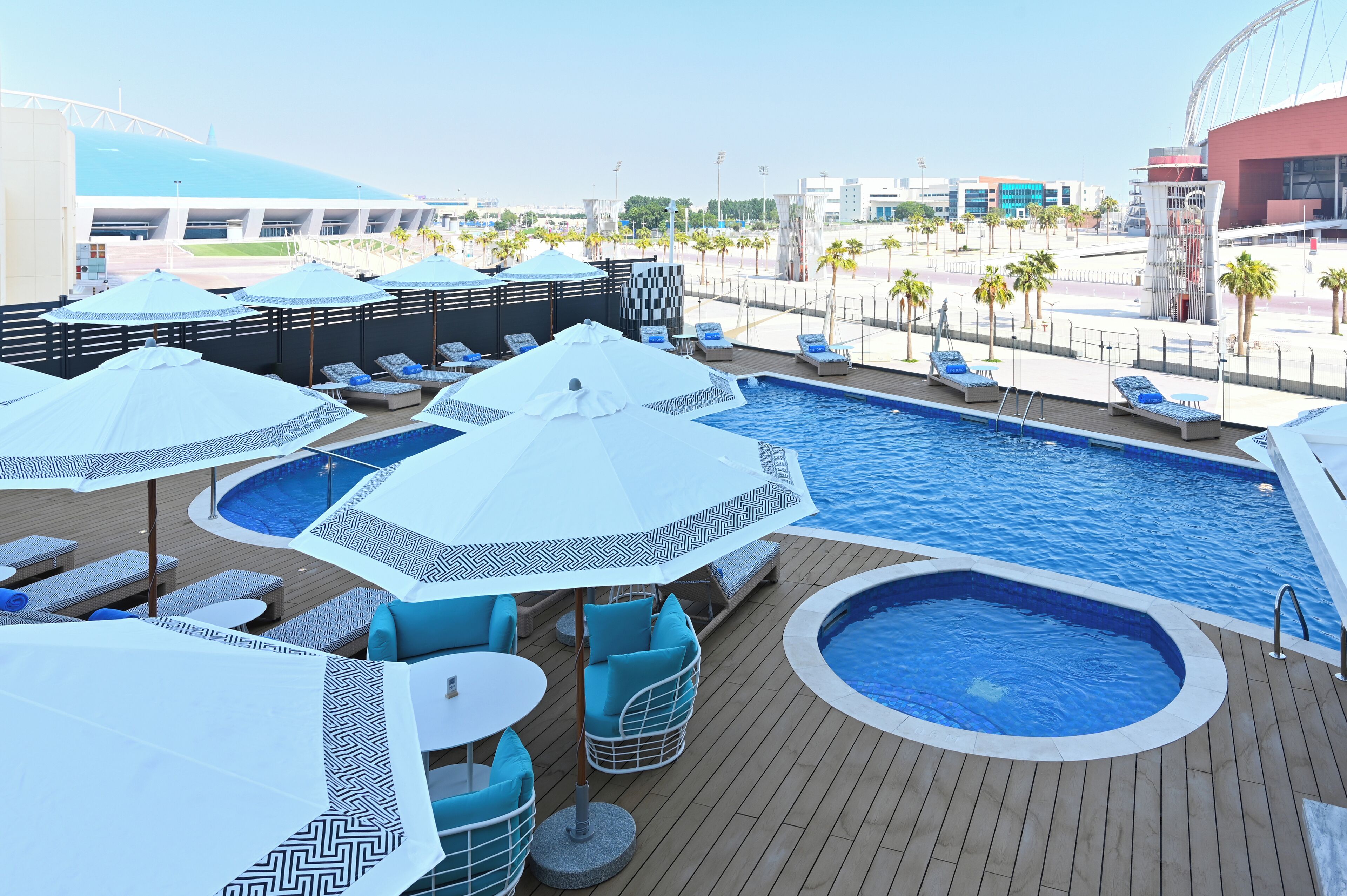 Un área de piscina en la azotea con múltiples sillas de descanso y sombrillas, un jacuzzi más pequeño y vistas de la ciudad circundante.