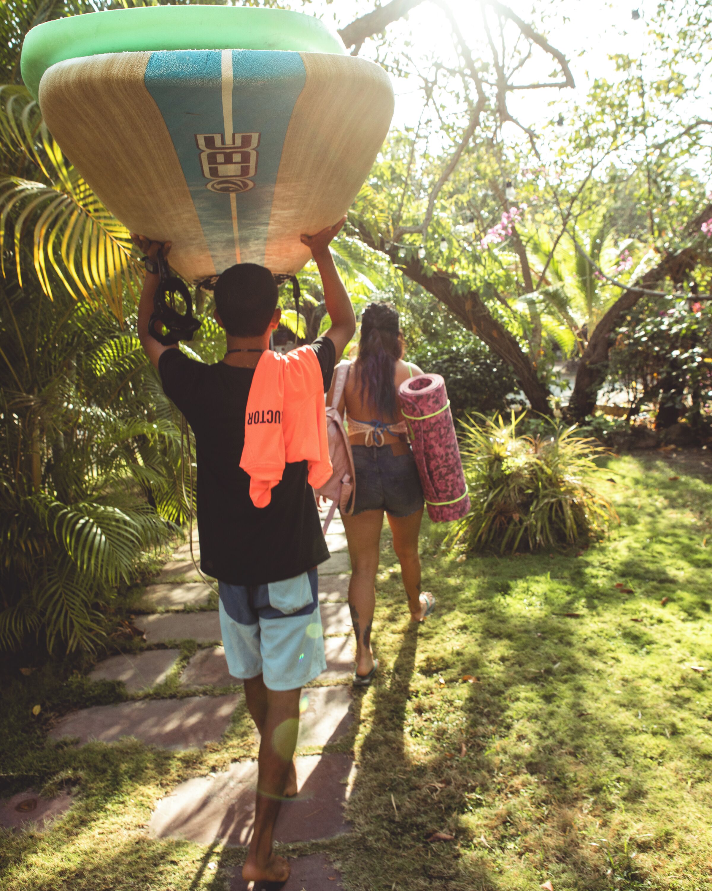 Two people carrying a surfboard on a path surrounded by greenery.