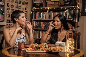 Dos personas cenando en una mesa con diversos platos, una botella y una guirnalda de luces al fondo.