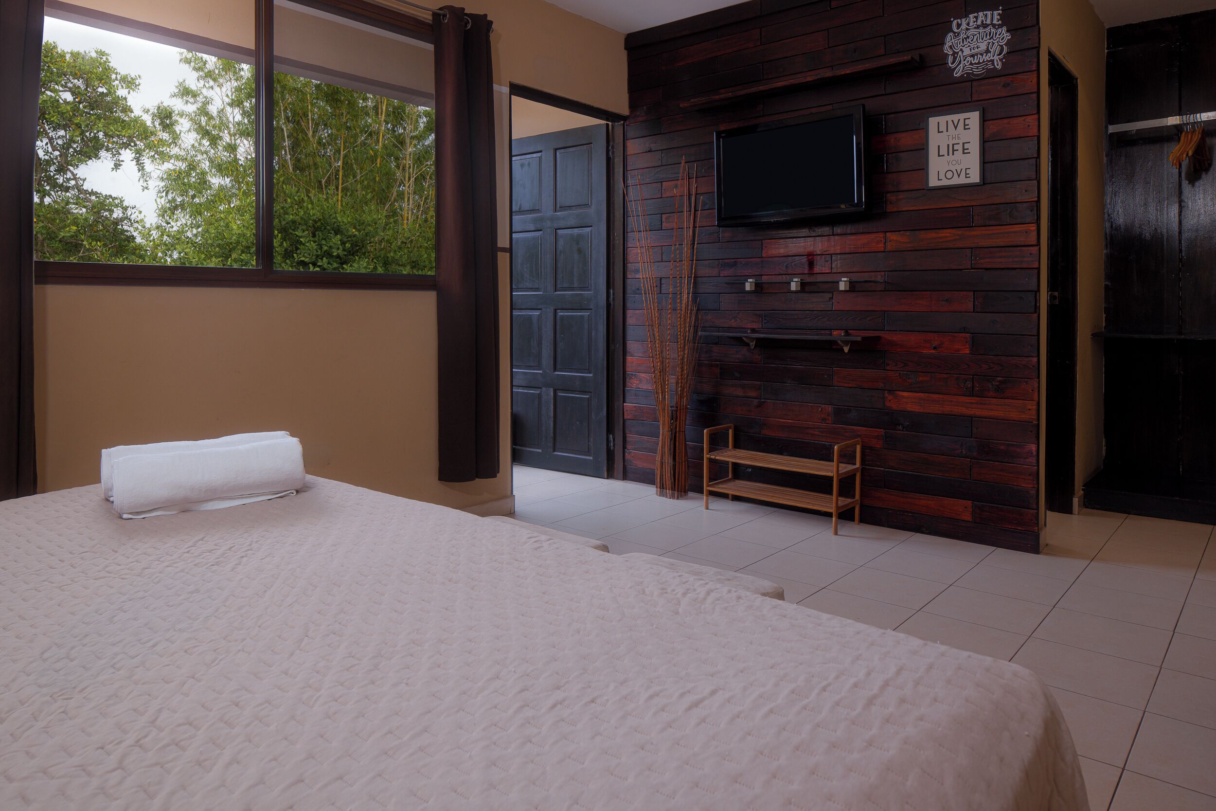 A bedroom with a large bed, a TV mounted on a wooden wall, and a view of the outdoors.