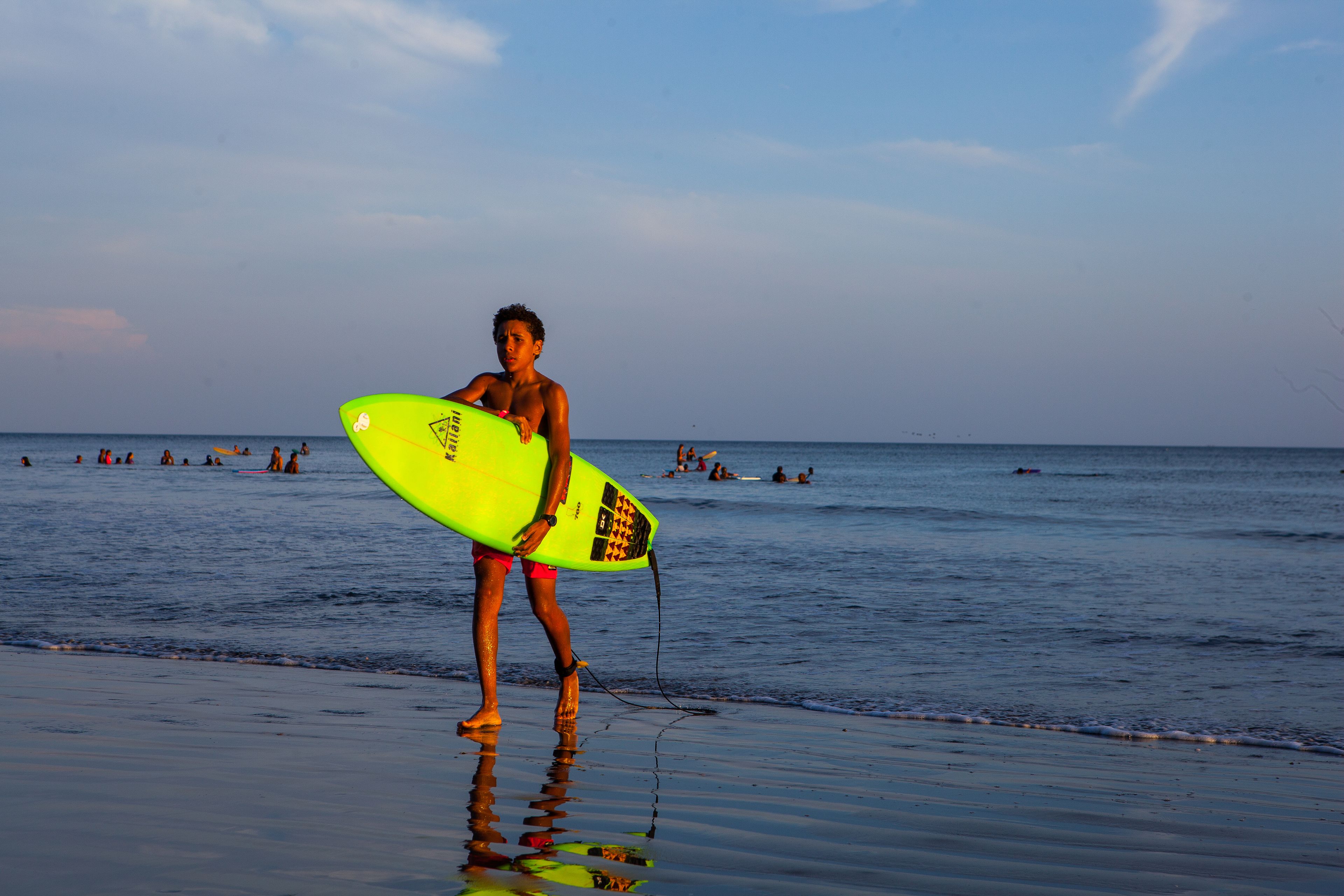 Iemand houdt een neongele surfplank vast op een strand, met mensen in het water en een heldere lucht.