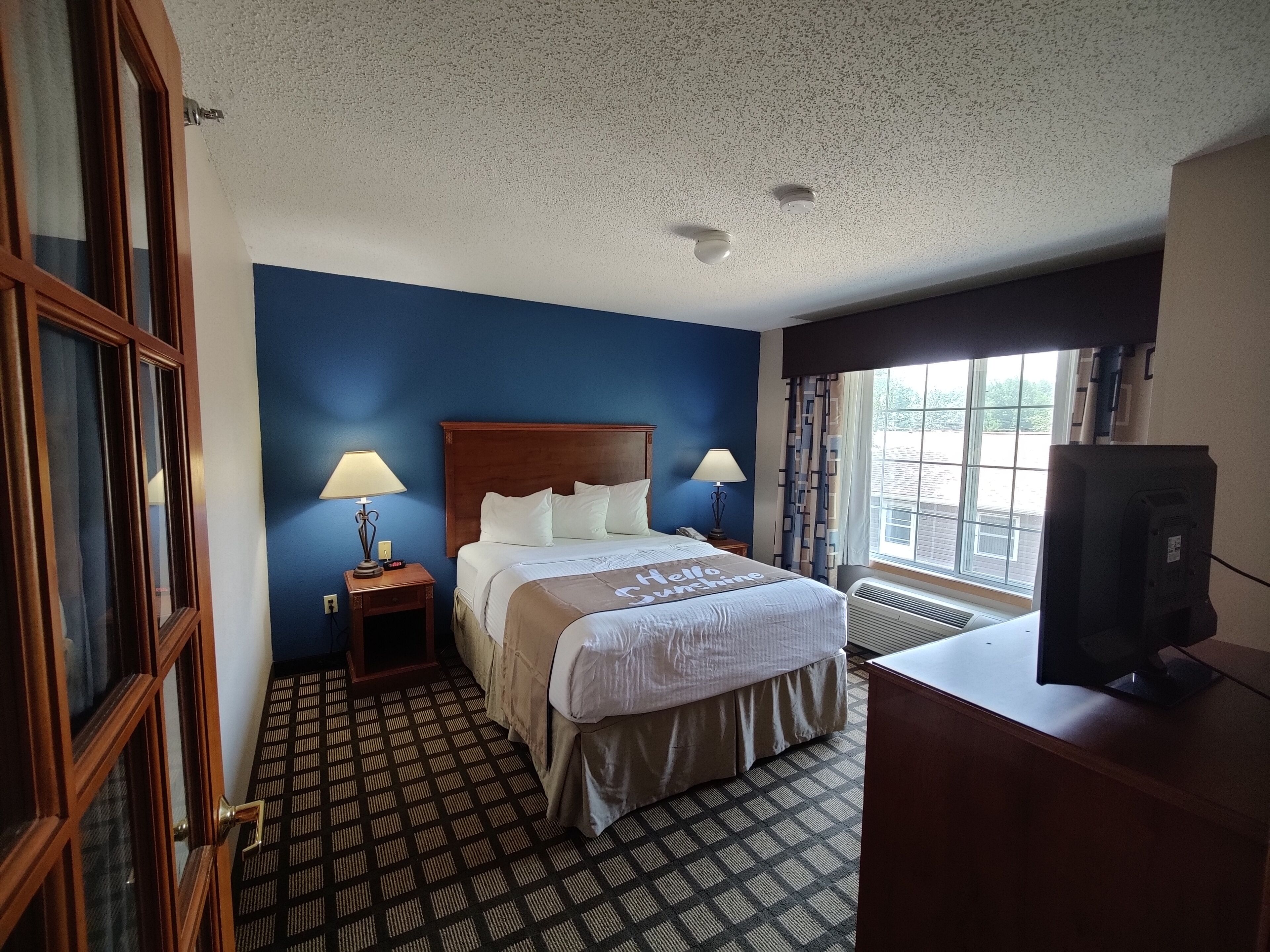 Habitación de hotel con cama, dos lámparas de noche, ventana con cortinas, televisor y alfombra estampada.